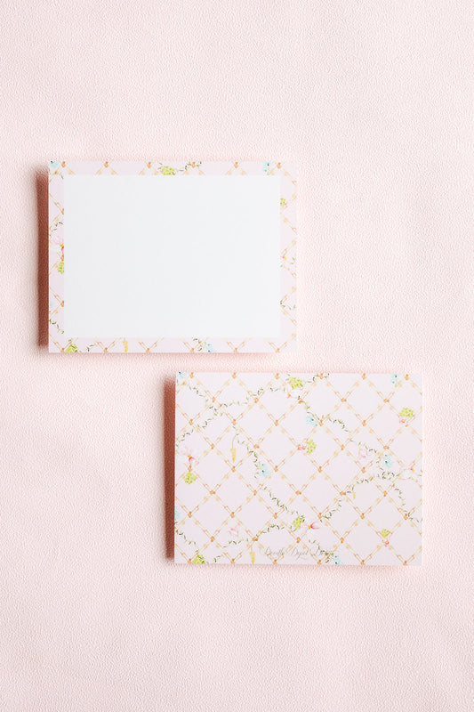 Two rectangular notepad sheets on a pale pink textured background. The top sheet has a white center with a delicate multicolor floral border.