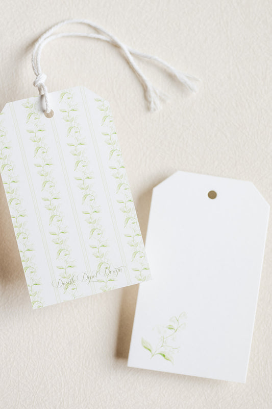 Two white gift tags with a small green floral print and white hanging strings, photographed on a light beige textured background.