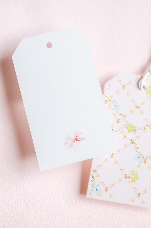 A white gift tag with a small pink flower illustration lies on a pale pink textured surface next to a white gift bag decorated with a gold lattice pattern and pastel floral details.