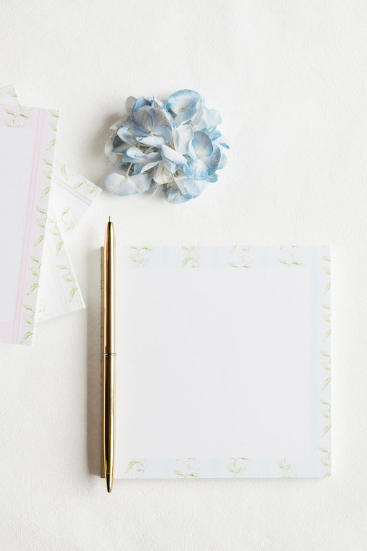 A flat-lay of a white notepad with a light blue and green floral border, a gold pen, and a blue paper flower on a white background.