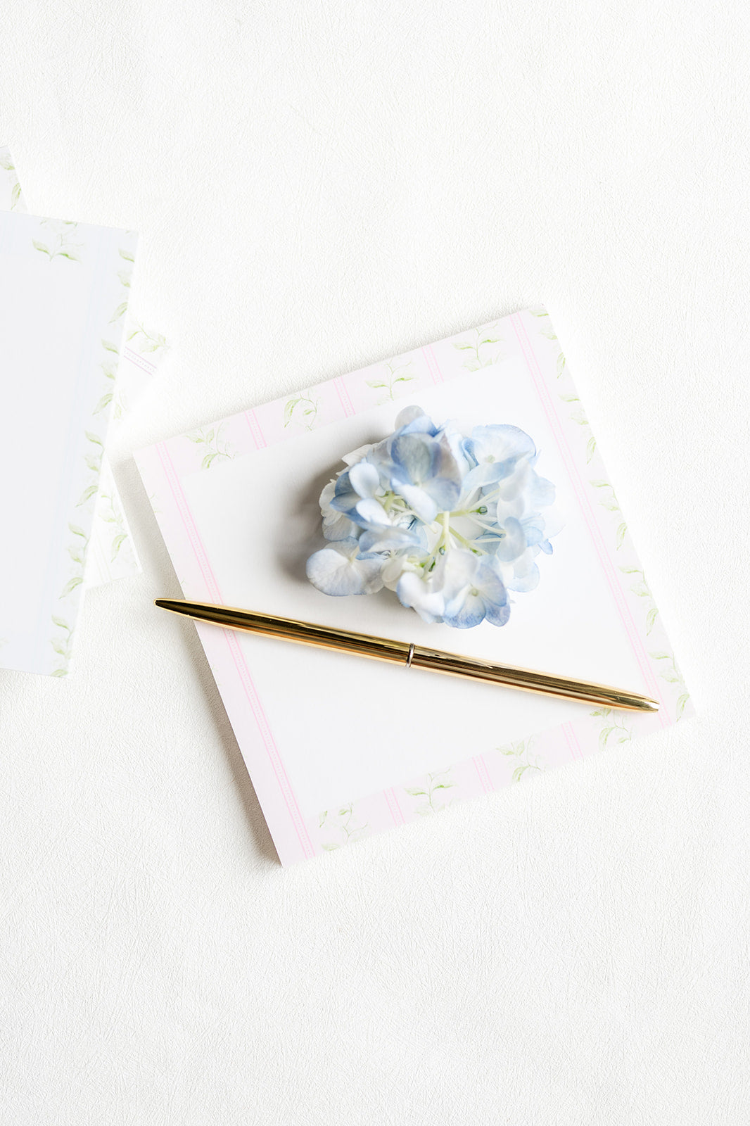 A gold metal pen resting on a square white notepad with a light pink border and a small blue hydrangea flower on top, photographed on a clean white surface.
