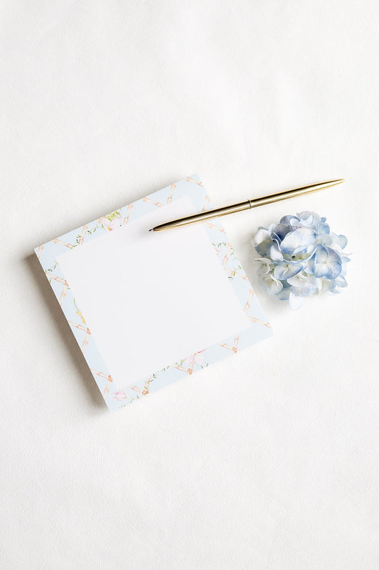 A square white notepad with a delicate pastel floral border, styled on a white background next to a gold pen and a small blue hydrangea flower.