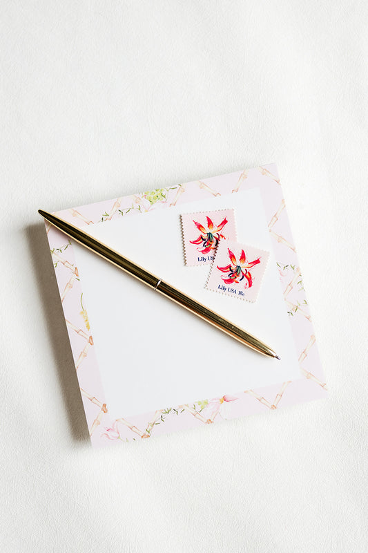 A gold pen resting diagonally on a square white notepad with a delicate gold foil border and two vintage-style postage stamps featuring red flowers.