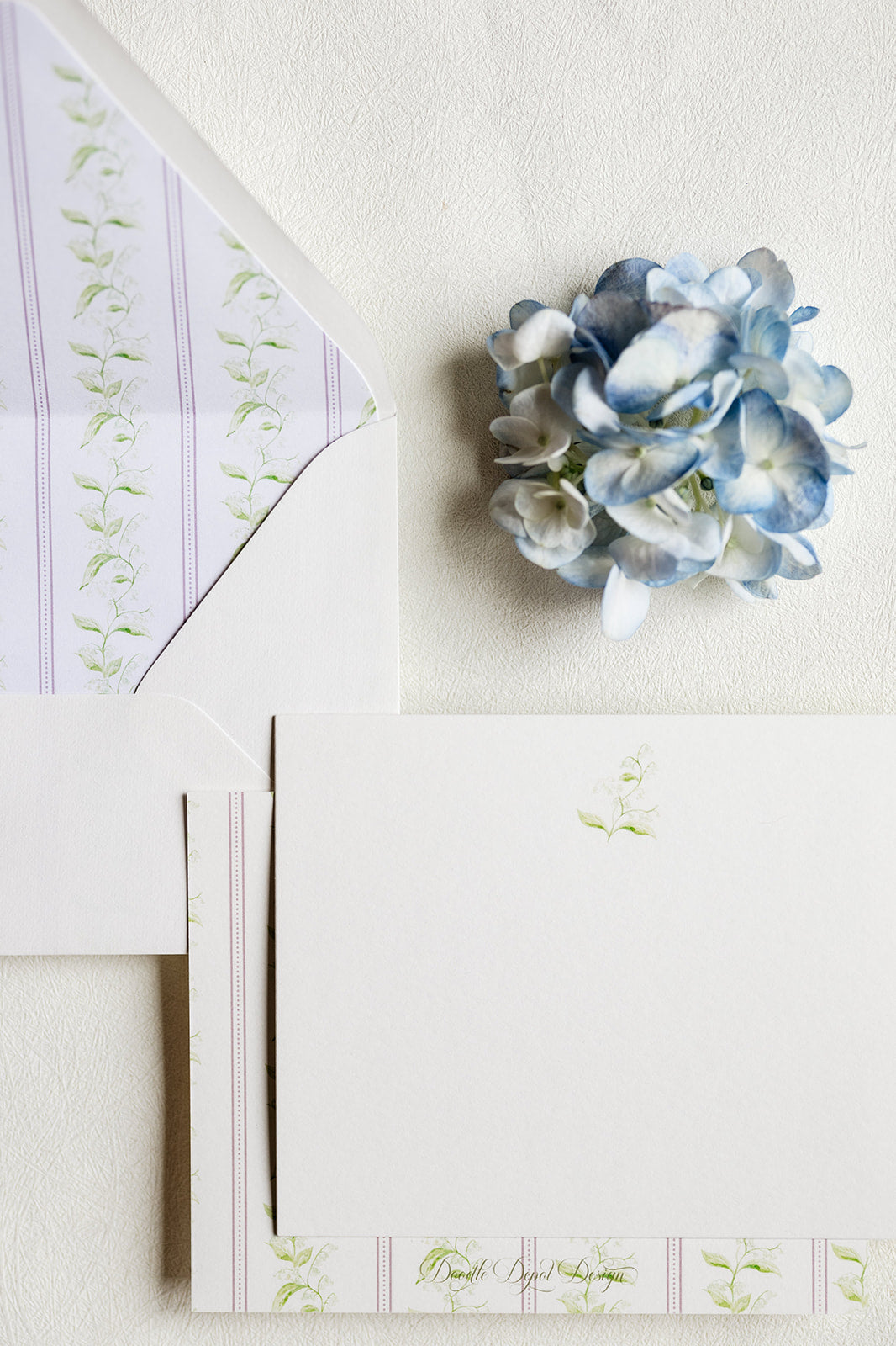 A styled flat-lay of white stationery with pale pink and green floral border details, an open white envelope, and a small blue hydrangea flower on a textured off-white background.