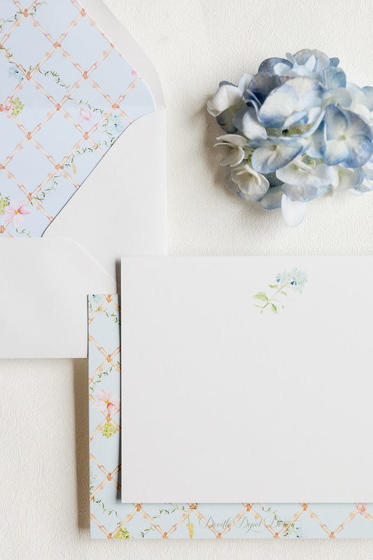 A flat-lay of a white stationery card with a small blue floral print, layered over patterned envelopes featuring a peach lattice design and blue hydrangea accents on a textured off-white background.