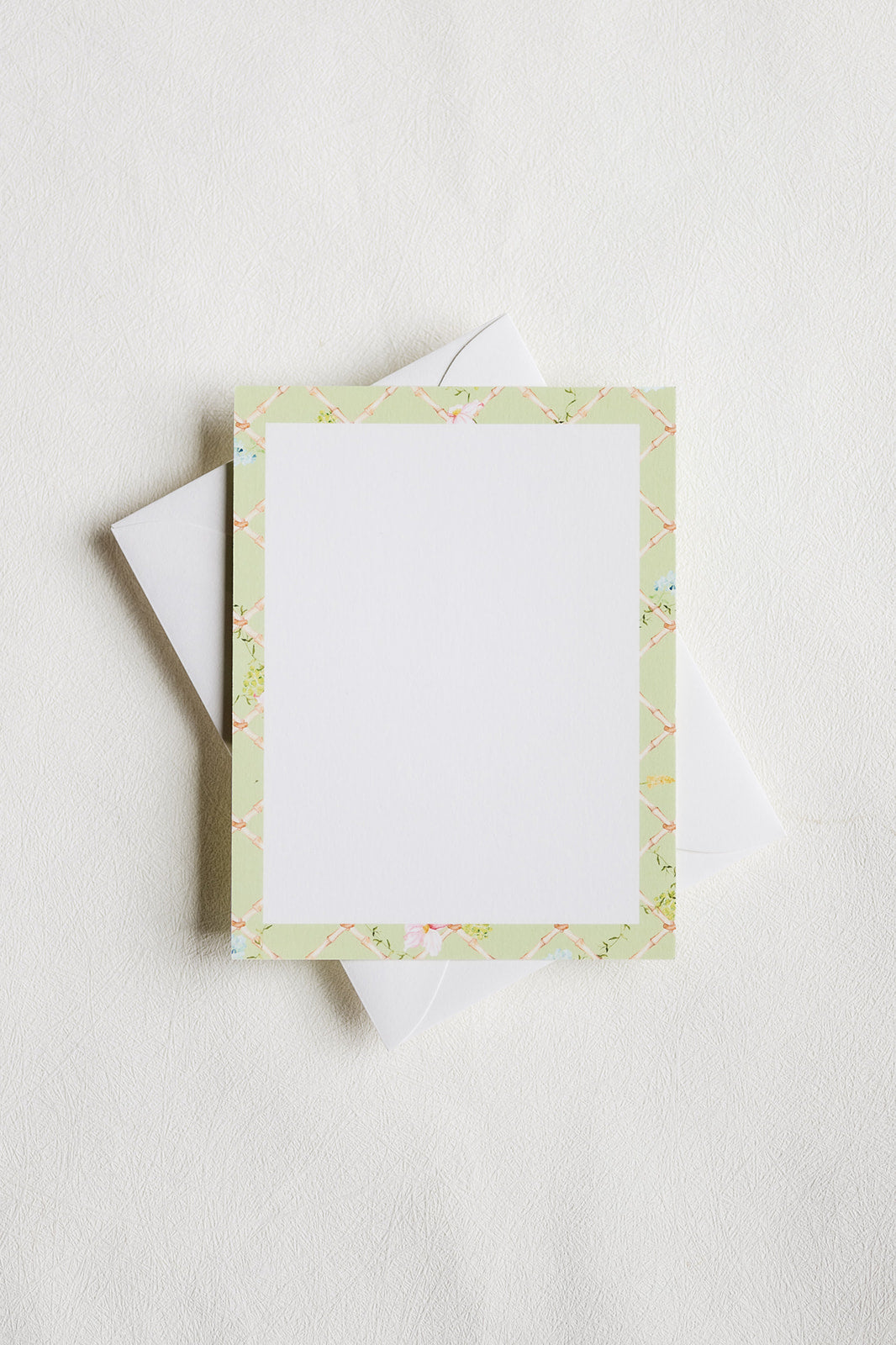 A square white writing card with a pale green floral border, shown with a white envelope on a light neutral background.
