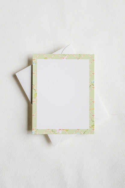 A square white writing card with a pale green floral border, shown with a white envelope on a light neutral background.