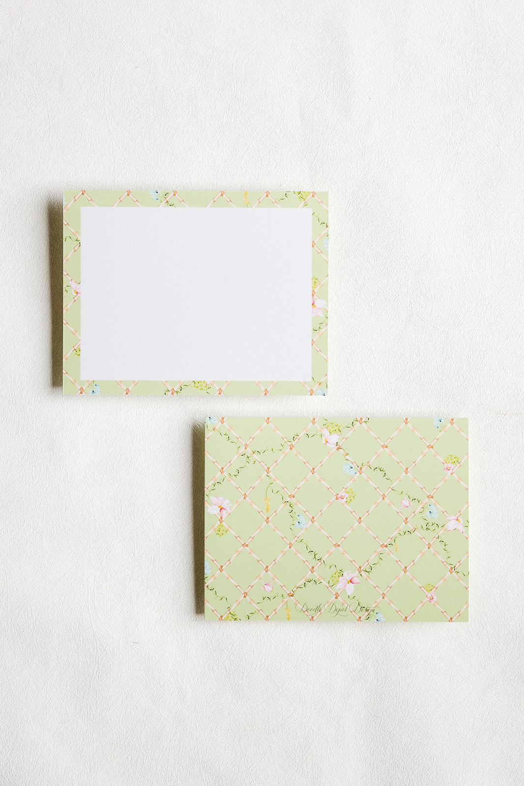 Two square paper notepads or cards with a pale green floral border pattern, one showing a blank white center and the other showing the patterned cover, arranged on a light textured background.