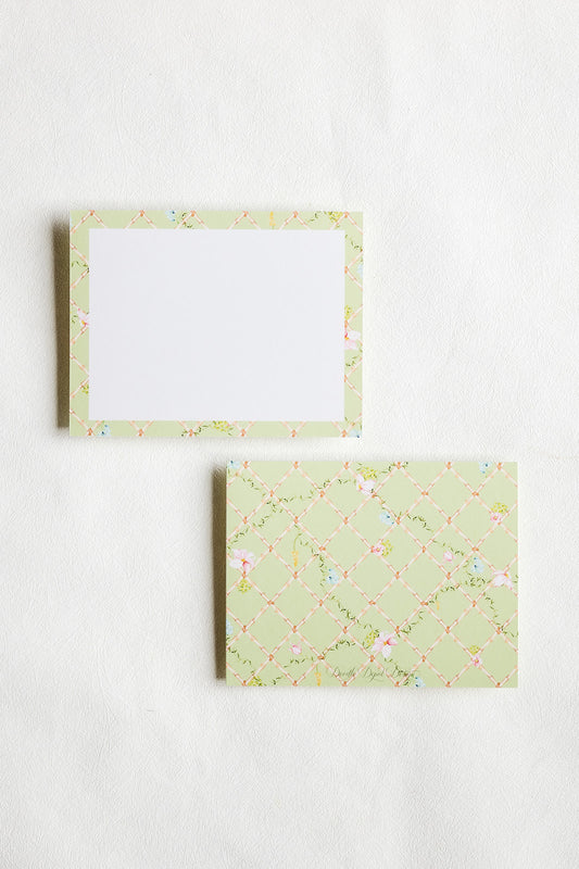 Two square paper notepads or cards with a pale green floral border pattern, one showing a blank white center and the other showing the patterned cover, arranged on a light textured background.
