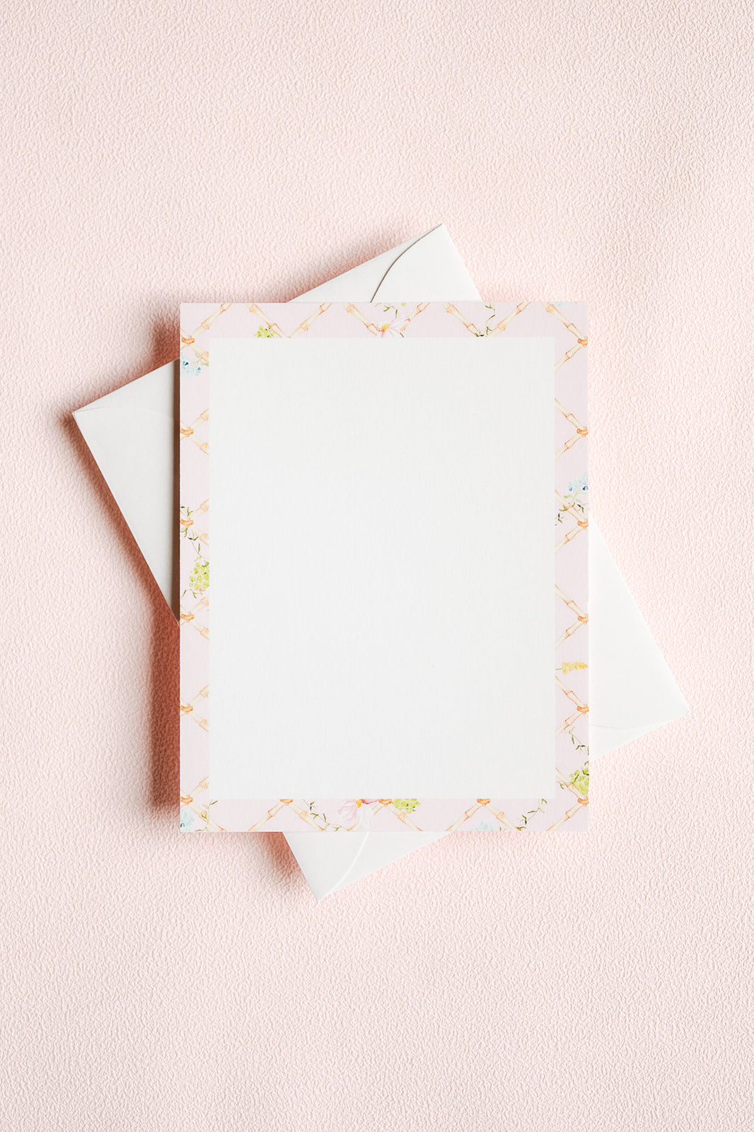 A flat-lay of a white stationery card with a delicate pastel border pattern, resting on top of white envelopes against a textured light pink background.