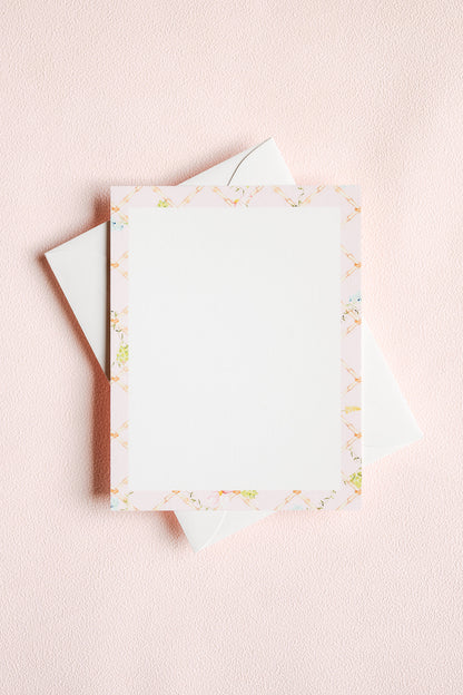 A flat-lay of a white stationery card with a delicate pastel border pattern, resting on top of white envelopes against a textured light pink background.