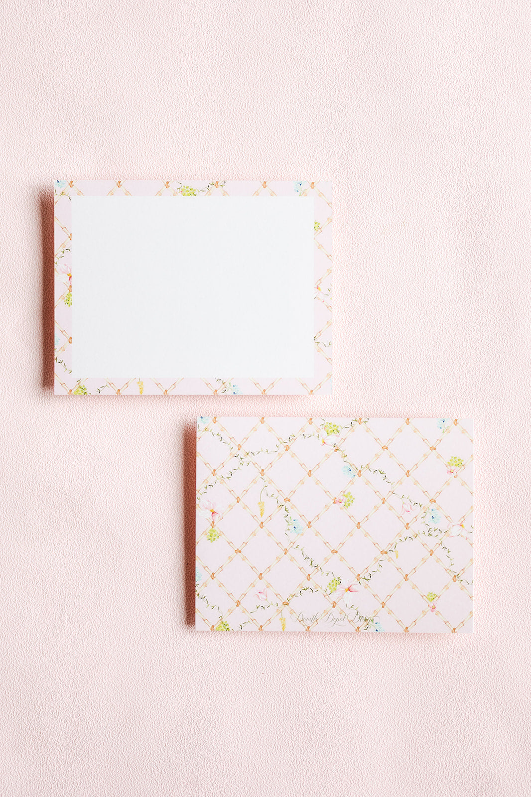 Two rectangular notepad sheets on a pale pink textured background. The top sheet has a white center with a delicate multicolor floral border.