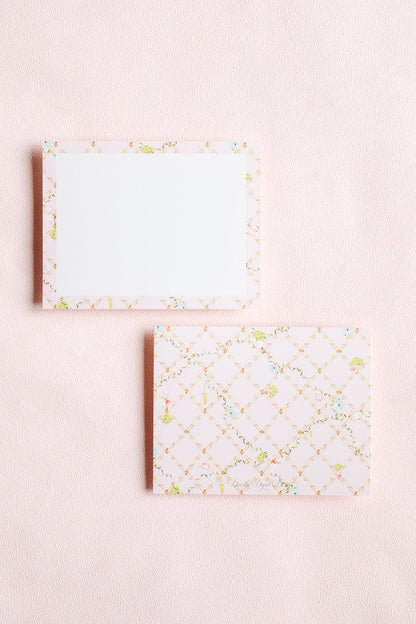 Two rectangular notepad sheets on a pale pink textured background. The top sheet has a white center with a delicate multicolor floral border.