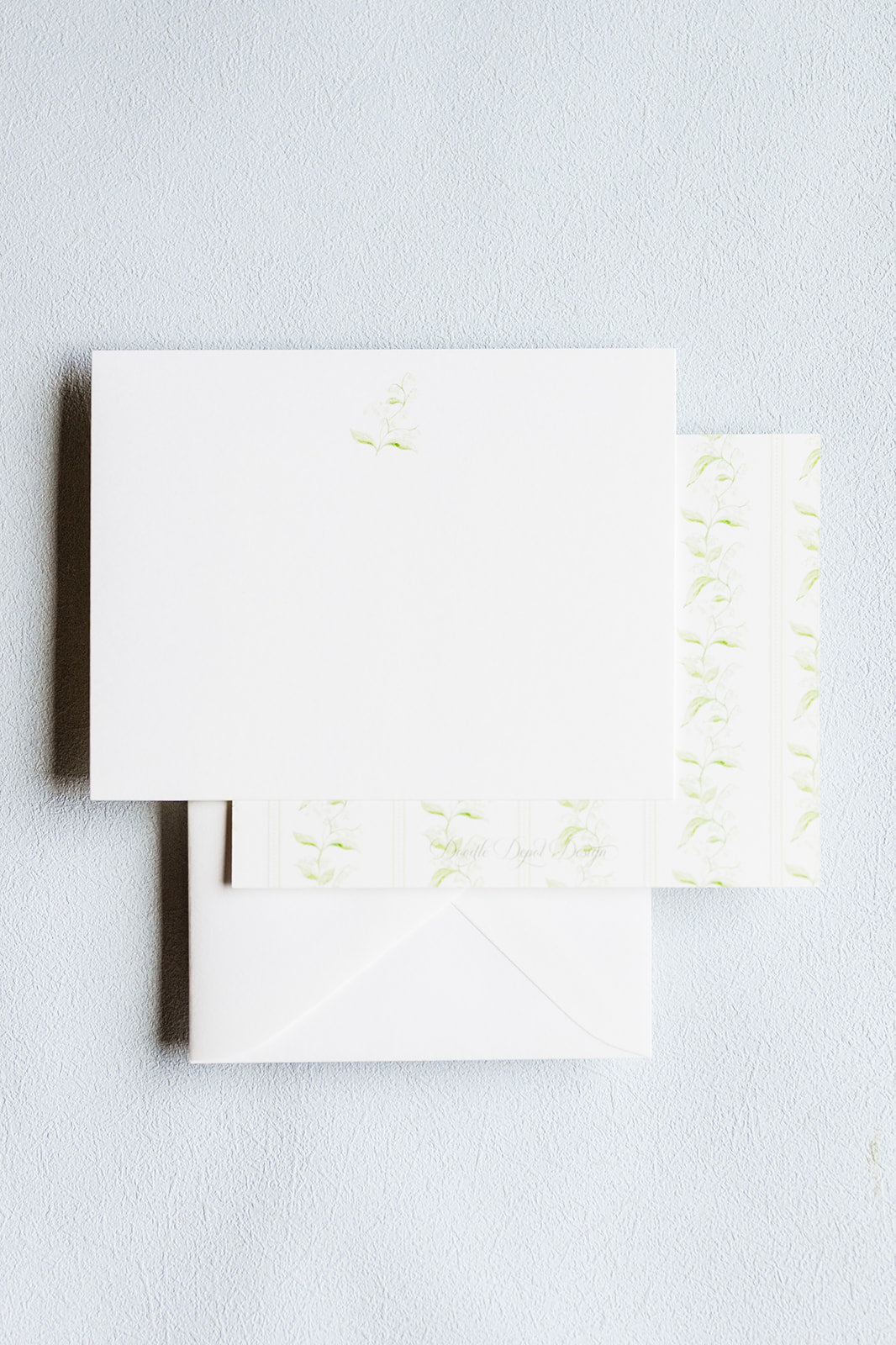 A stack of white stationery cards and envelopes with delicate pale green floral embroidery, photographed on a light gray textured background.