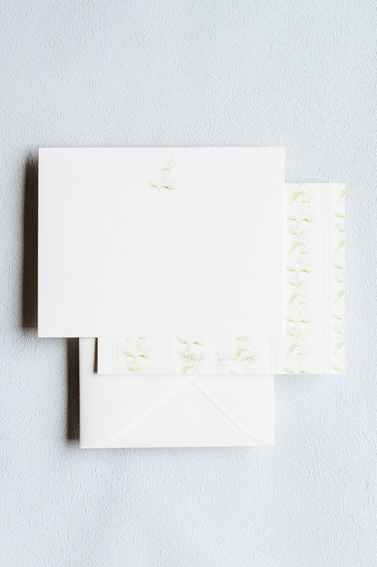 A stack of white stationery cards and envelopes with delicate pale green floral embroidery, photographed on a light gray textured background.