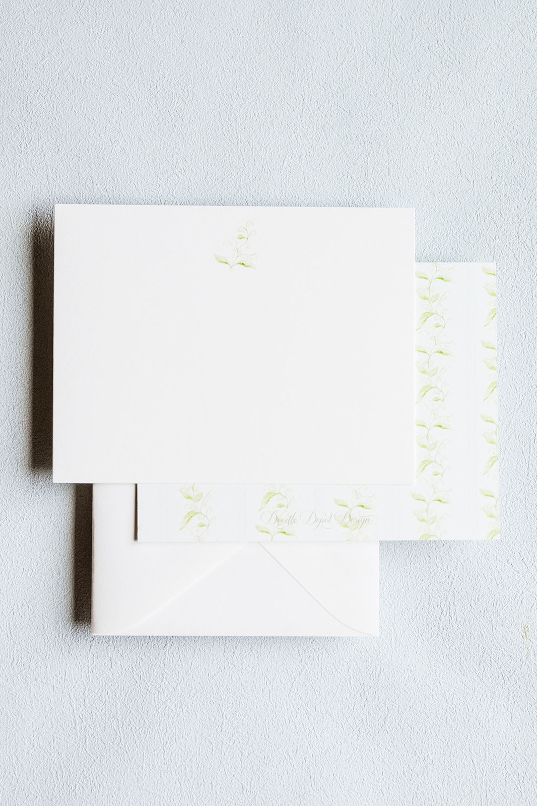White stationery set with a square card, matching envelope, and a narrow strip of patterned paper featuring light green floral motifs, arranged on a pale textured background.