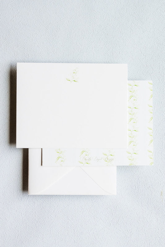 White stationery set with a square card, matching envelope, and a narrow strip of patterned paper featuring light green floral motifs, arranged on a pale textured background.