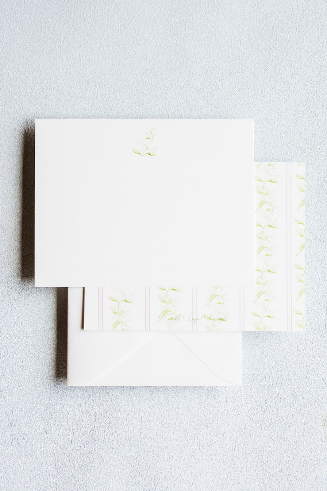 White stationery set with a square card, envelope, and matching patterned paper featuring light green floral and scroll motifs, arranged on a light gray textured background.