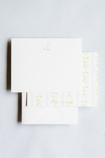 White stationery set with a square card, envelope, and matching patterned paper featuring light green floral and scroll motifs, arranged on a light gray textured background.