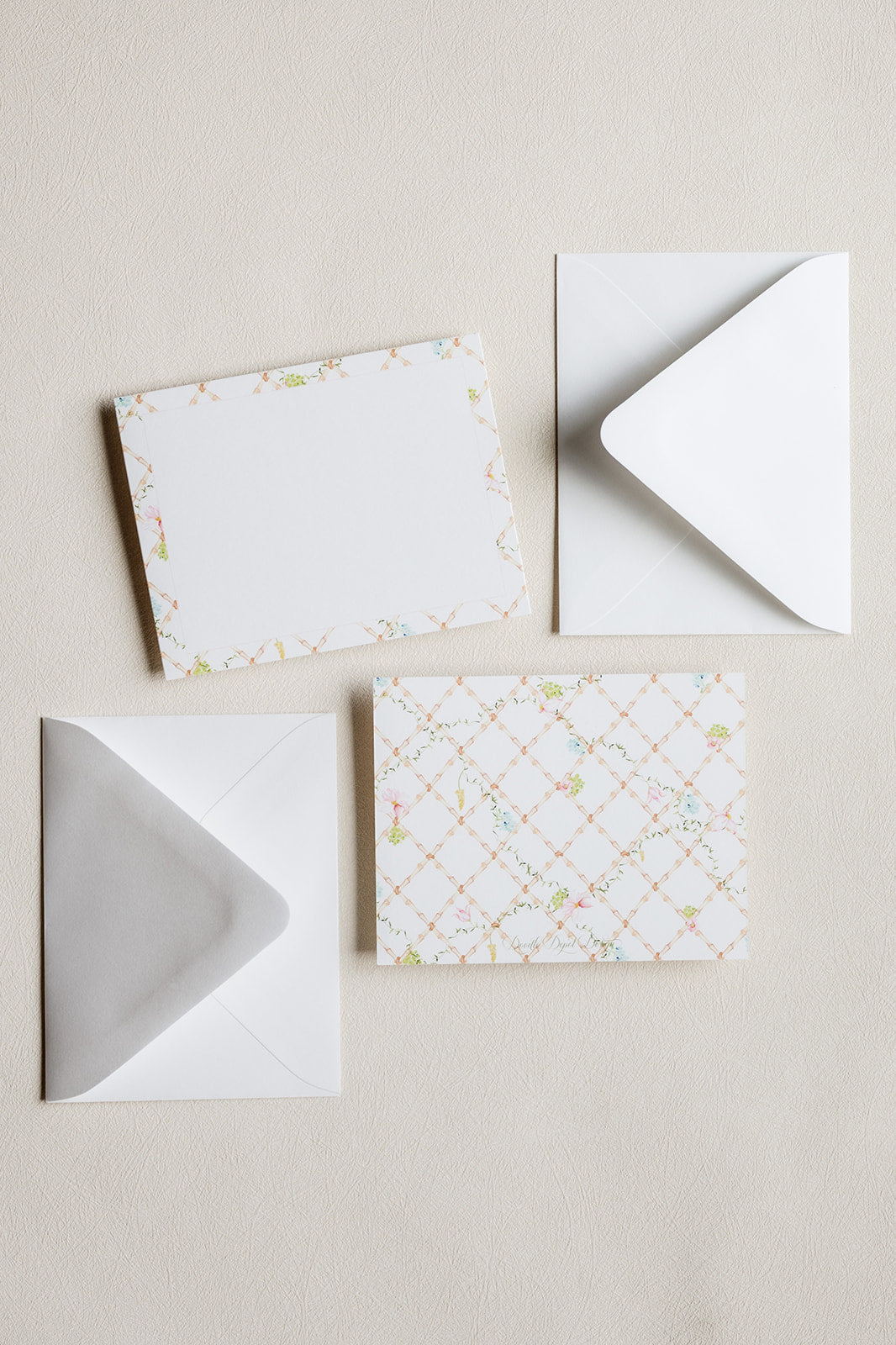 A set of white stationery pieces arranged on a light beige textured background, including a patterned card with a delicate floral border, a white card, and two white envelopes.