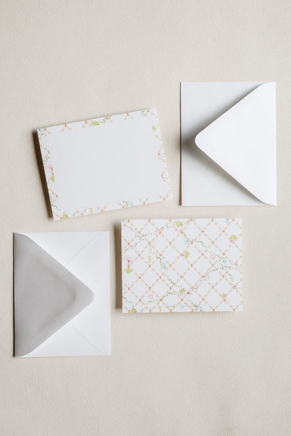 A set of white stationery pieces arranged on a light beige textured background, including a patterned card with a delicate floral border, a white card, and two white envelopes.