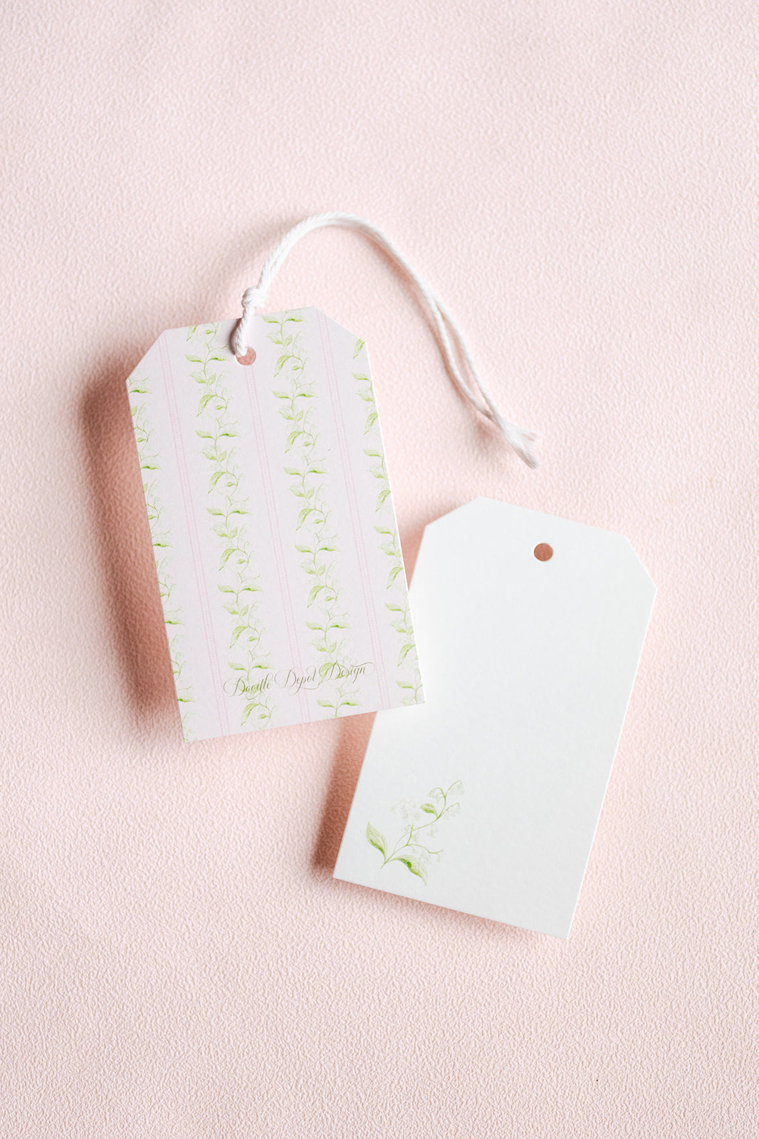 Two decorative gift tags with a white string, one featuring pale green floral stripes and the other plain white, set against a soft pink textured background.
