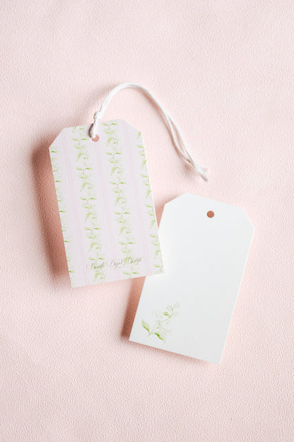 Two decorative gift tags with a white string, one featuring pale green floral stripes and the other plain white, set against a soft pink textured background.