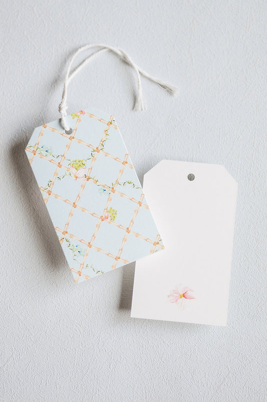 Two decorative gift tags on a light gray textured background. One tag has a pale blue background with a peach lattice pattern and small floral details, attached to a white string.