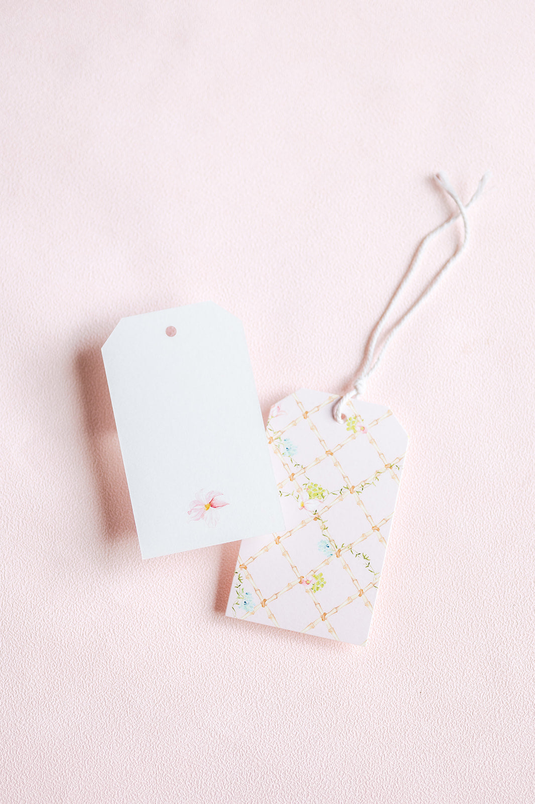 Two decorative gift tags on a soft pink background. One tag is plain white with a small pink floral detail, and the other has a white background with a delicate gold lattice pattern and small pastel floral accents.