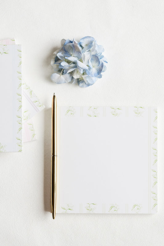 A gold pen lies on a white notepad with a light green floral border, arranged on a white surface next to a small blue hydrangea flower and part of another matching paper sheet.