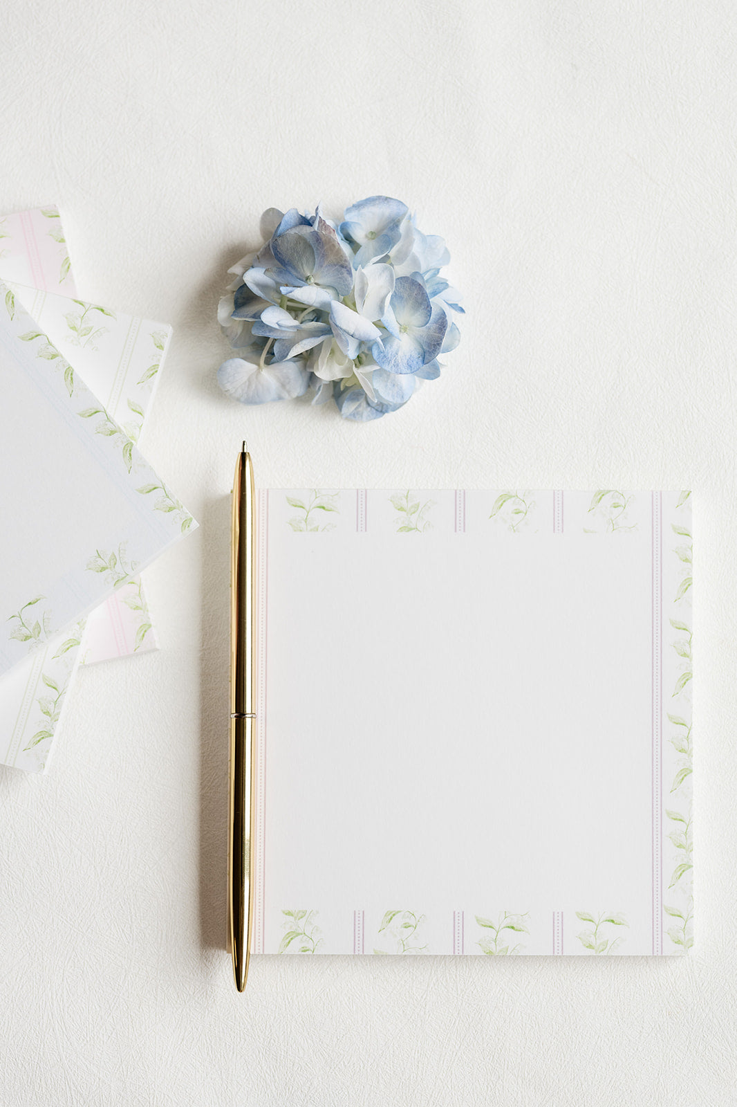 A flat-lay of a white notepad with a delicate pale green floral border, accompanied by a gold pen and a light blue hydrangea on a textured white background.
