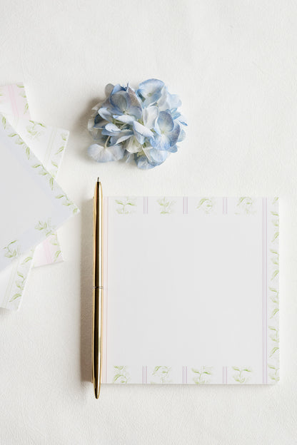 A flat-lay of a white notepad with a delicate pale green floral border, accompanied by a gold pen and a light blue hydrangea on a textured white background.