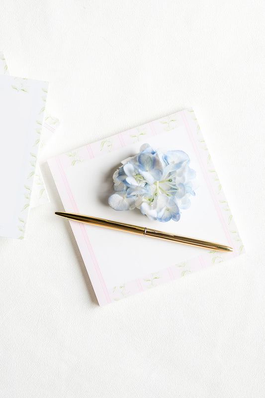 A gold metal pen resting on a square white notepad with a light pink border and a small blue hydrangea flower on top, photographed on a clean white surface.