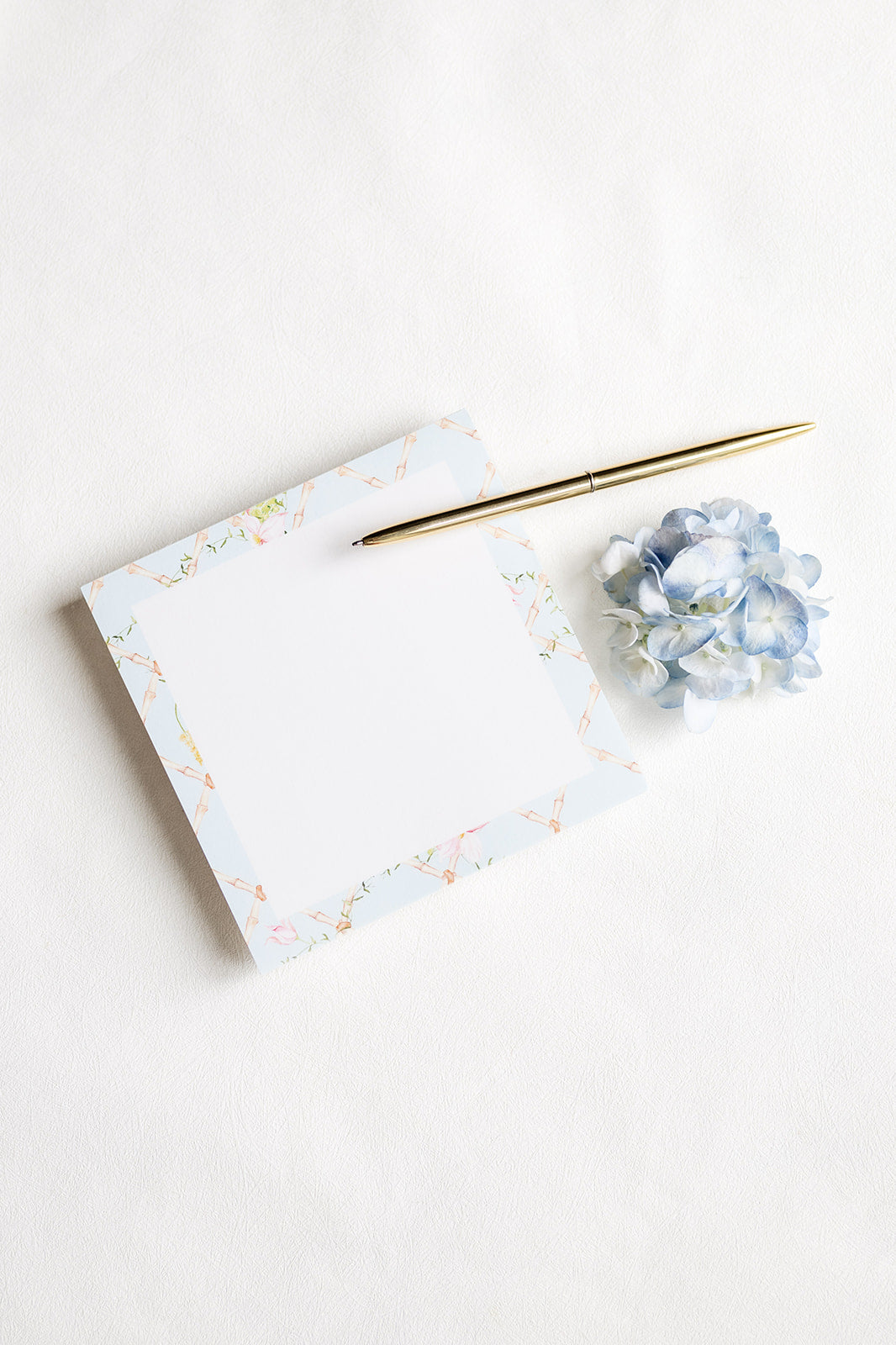 A square white notepad with a delicate pastel floral border, styled on a white background next to a gold pen and a small blue hydrangea flower.