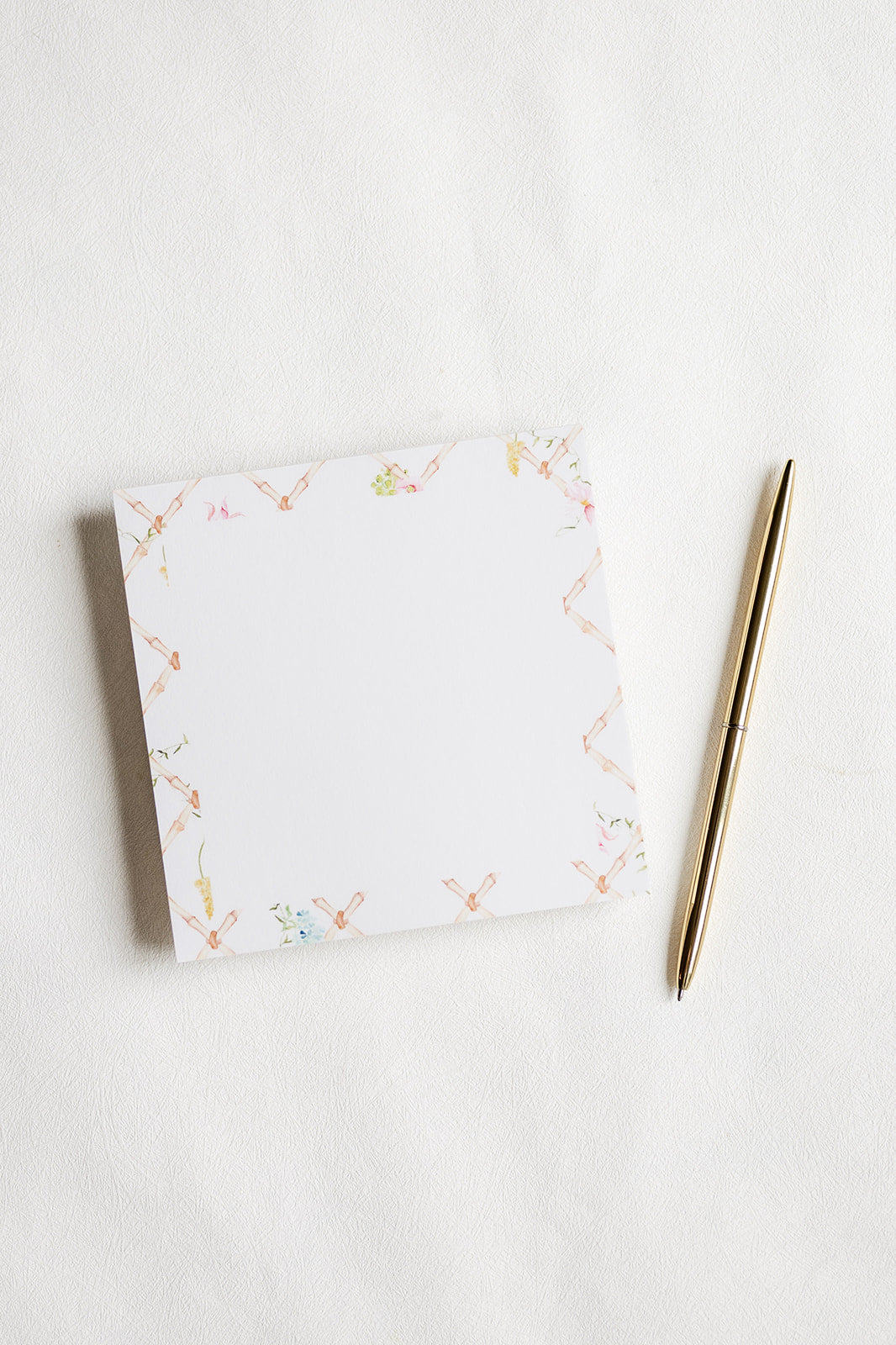 A square white notepad with a delicate pastel floral border, placed on a white textured surface next to a slim gold pen.