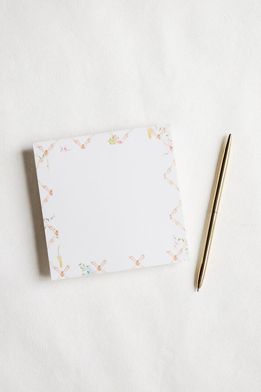 A square white notepad with a delicate pastel floral border, placed on a white textured surface next to a slim gold pen.