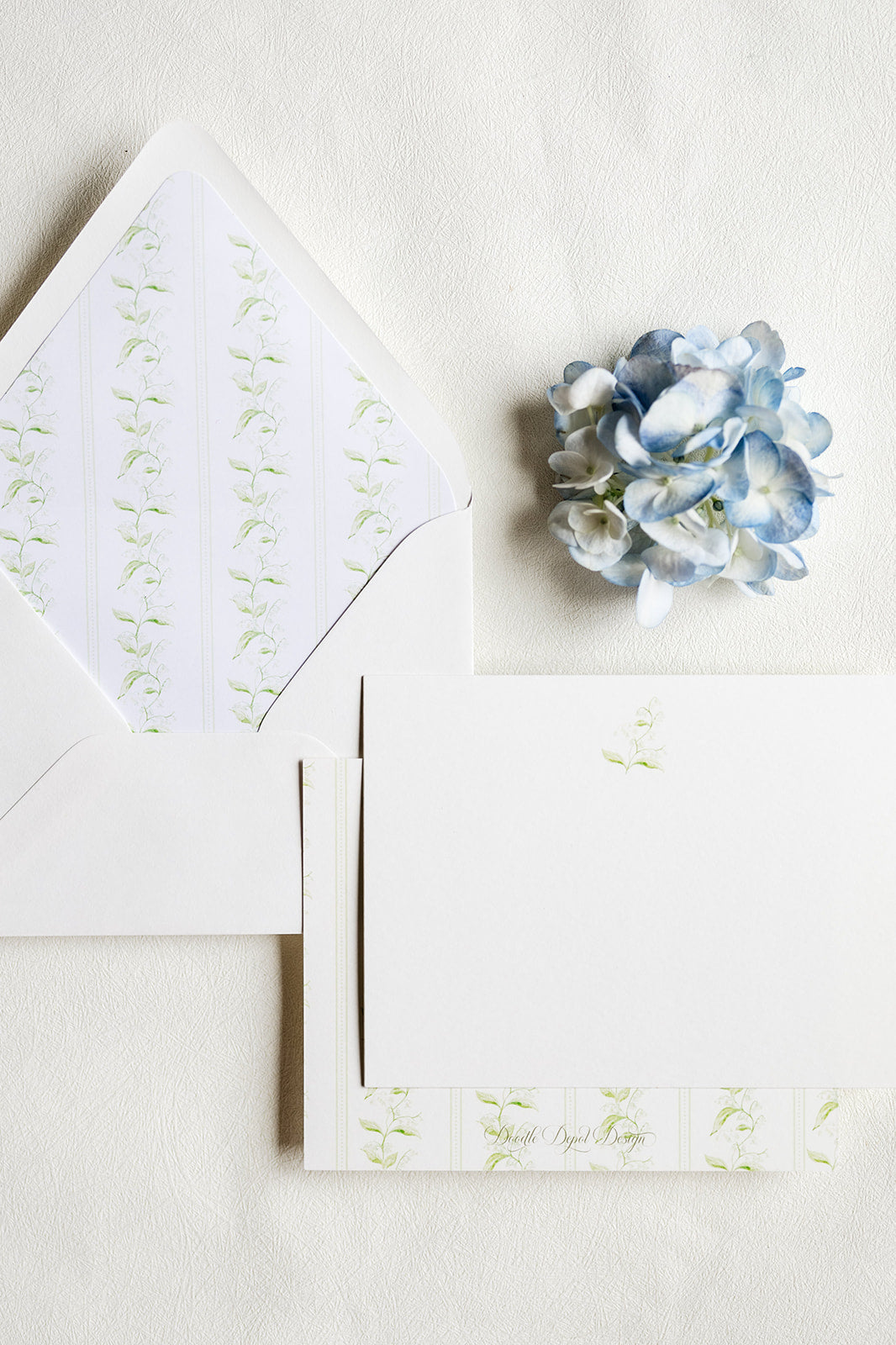 White stationery set with a floral envelope liner, matching envelope, folded card, and a small blue hydrangea flower on a textured white background.