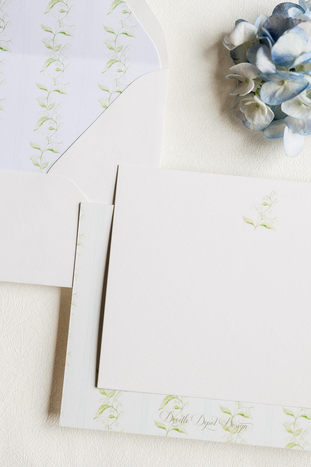 A styled flat-lay of white stationery with delicate light green floral vine prints, including a card, envelope, and matching paper, arranged on a textured cream surface next to a blue hydrangea.