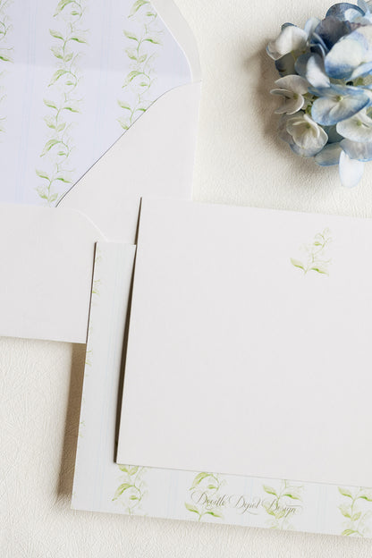 A styled flat-lay of white stationery with delicate light green floral vine prints, including a card, envelope, and matching paper, arranged on a textured cream surface next to a blue hydrangea.