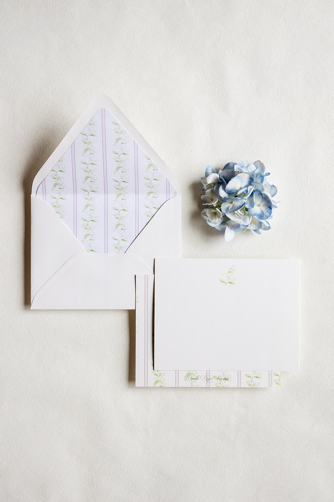 A flat-lay of white stationery on a textured off-white background, featuring an open envelope with a pale lavender and green floral patterned liner, a matching folded card with a small green floral print, and a small blue hydrangea flower.