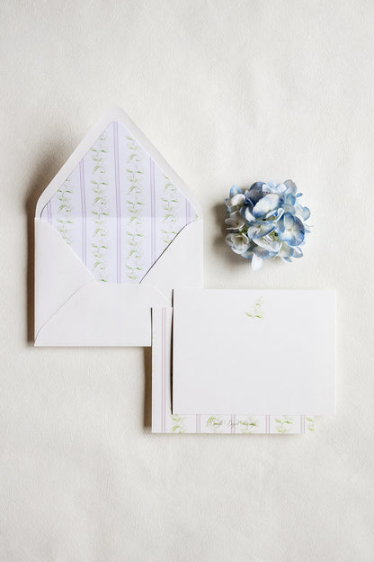 A flat-lay of white stationery on a textured off-white background, featuring an open envelope with a pale lavender and green floral patterned liner, a matching folded card with a small green floral print, and a small blue hydrangea flower.