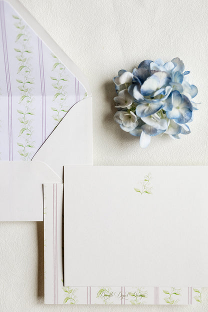 A styled flat-lay of white stationery with pale pink and green floral border details, an open white envelope, and a small blue hydrangea flower on a textured off-white background.