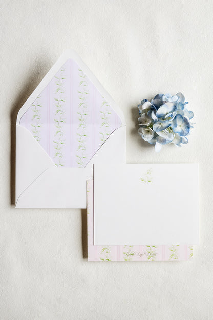 A stationery set featuring a white envelope with a pale lavender and green striped floral patterned liner, a matching white card with a small green floral print, and a decorative blue hydrangea flower on a textured off-white background.