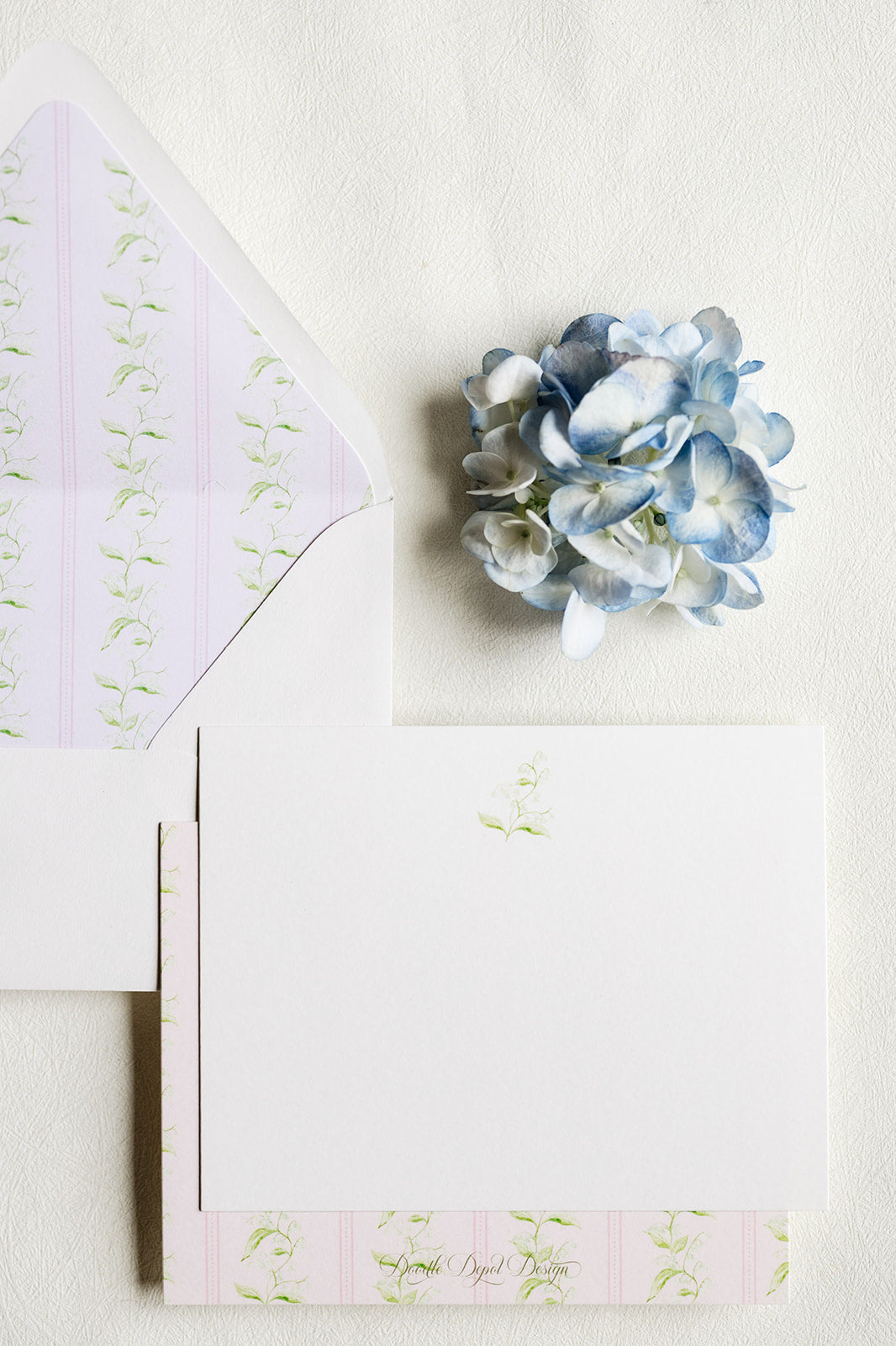 A styled flat-lay of white stationery on a textured off-white background, featuring a white card with a small green floral motif, a white envelope with pale pink and green striped patterned liner, and a decorative blue hydrangea flower.