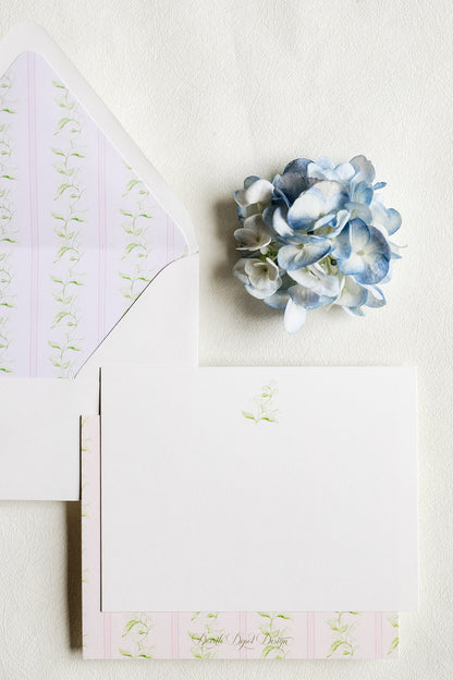 A styled flat-lay of white stationery on a textured off-white background, featuring a white card with a small green floral motif, a white envelope with pale pink and green striped patterned liner, and a decorative blue hydrangea flower.
