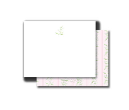 A white rectangular card or envelope with a small pale green floral print near the top, layered over a pink patterned sheet with repeating green botanical motifs, shown on a plain white background.