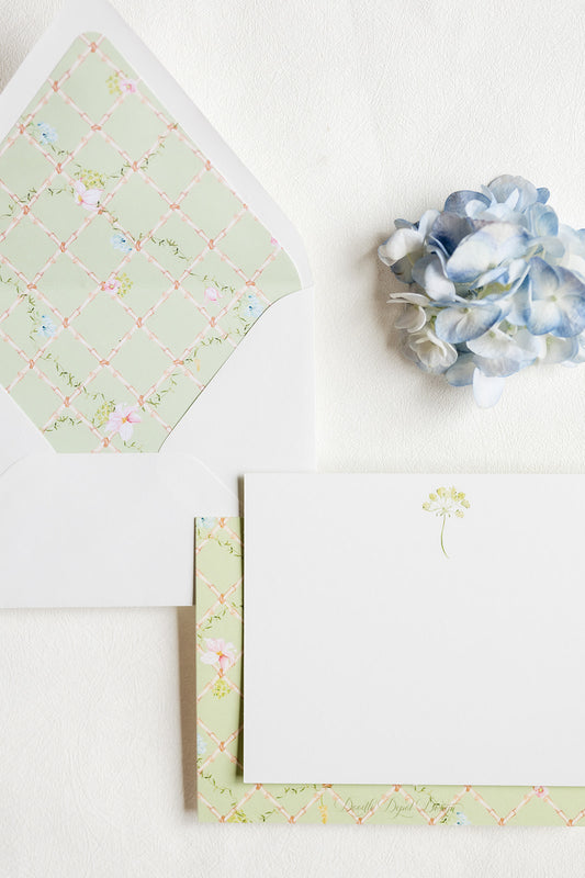 A styled flat-lay of white stationery with a pale green floral lattice patterned envelope and matching card border, plus a small blue hydrangea flower on a white background.
