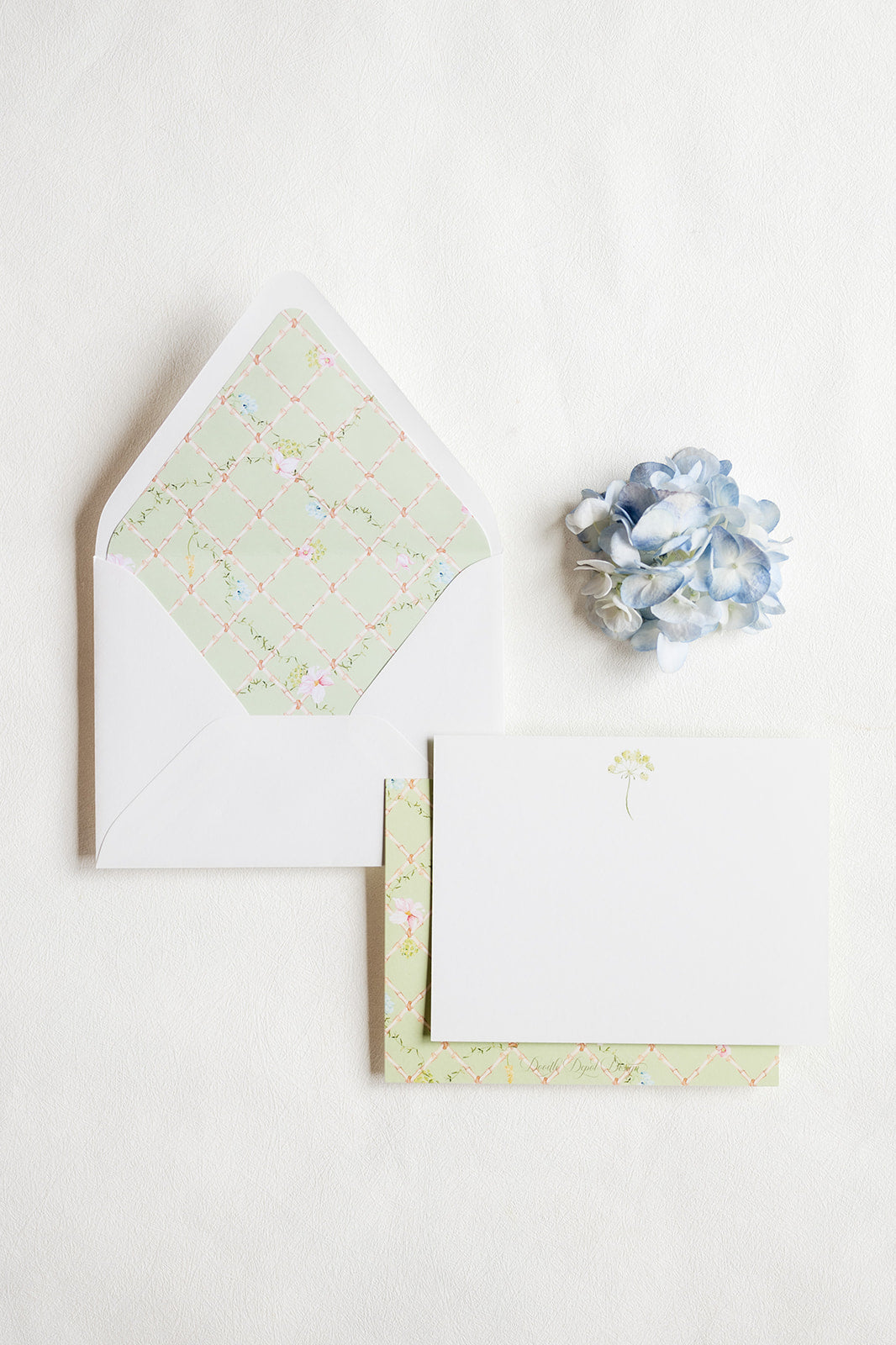 A styled flat-lay of stationery on a white background, featuring a white envelope with a pale green floral patterned inner flap, a white card with a small green umbrella illustration, patterned paper sheets, and a small blue hydrangea flower.