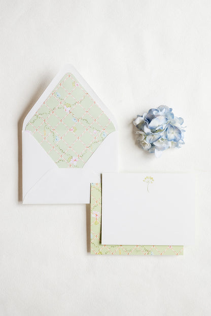 A styled flat-lay of stationery on a white background, featuring a white envelope with a pale green floral patterned inner flap, a white card with a small green umbrella illustration, patterned paper sheets, and a small blue hydrangea flower.