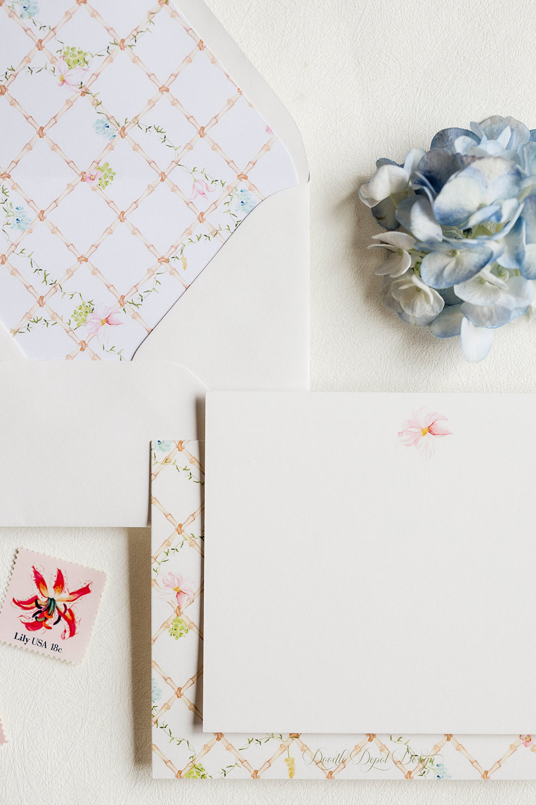 A styled flat-lay of white stationery with delicate pastel floral and lattice-patterned paper, a blue hydrangea flower, and a small decorative stamp on a textured off-white background.